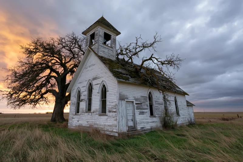 Abandoned white clapboard church with gothic arched windows and bell tower in a rural field at sunset