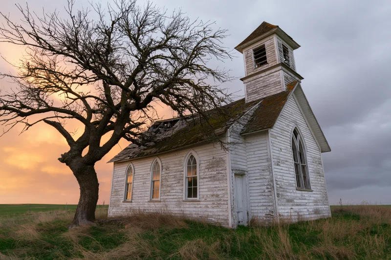 Abandoned white clapboard church with gothic arched windows and steeple in a rural field at sunset
