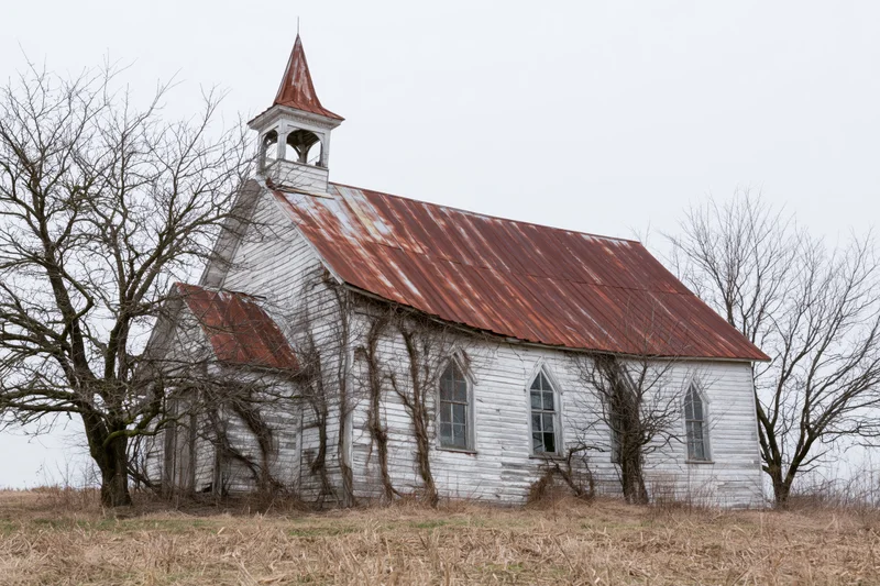 Abandoned white clapboard church with rusted corrugated roof and overgrown vines in a rural field