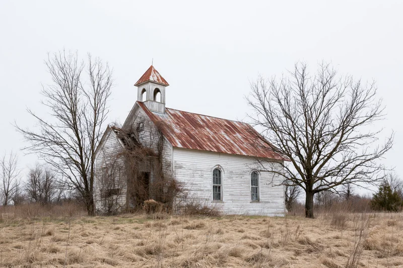 Abandoned white clapboard church with rusted corrugated roof in a rural field surrounded by bare trees