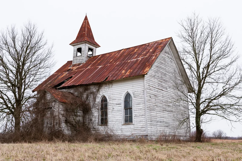 Abandoned white clapboard church with rusted corrugated roof in rural field surrounded by bare trees
