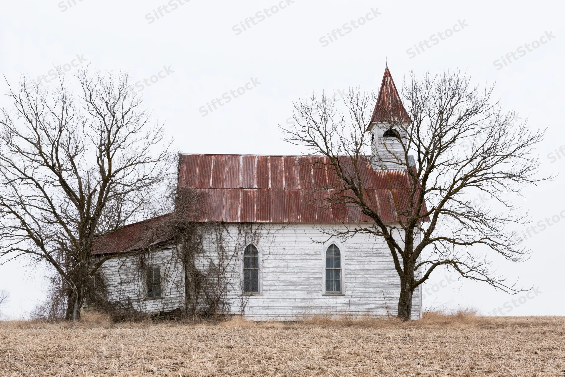 Abandoned white clapboard church with rusted corrugated roof in a dry field under overcast sky
