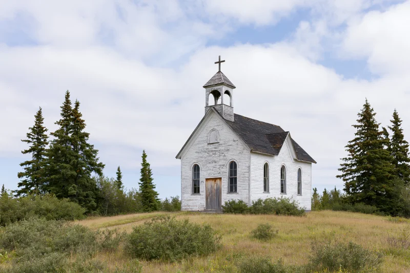 Small white clapboard church with bell steeple and cross standing in a grassy field surrounded by evergreen trees under a cloudy sky