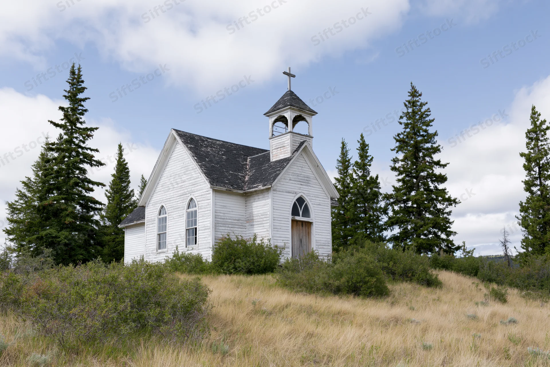 Small white clapboard church with bell steeple and cross in rural field surrounded by evergreen trees under blue sky