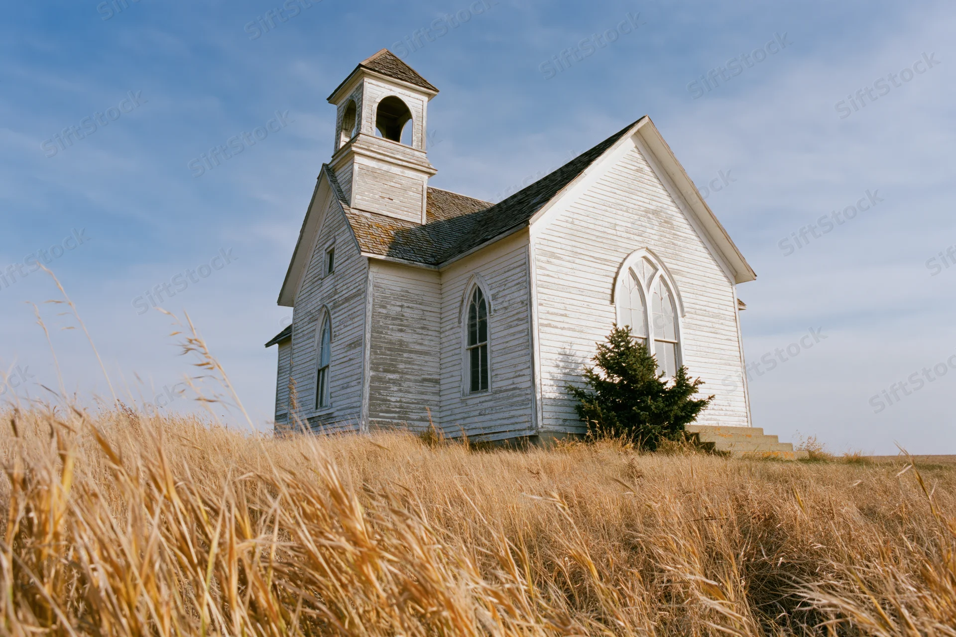 Abandoned white clapboard church with small bell tower in a golden grassy prairie field under blue sky