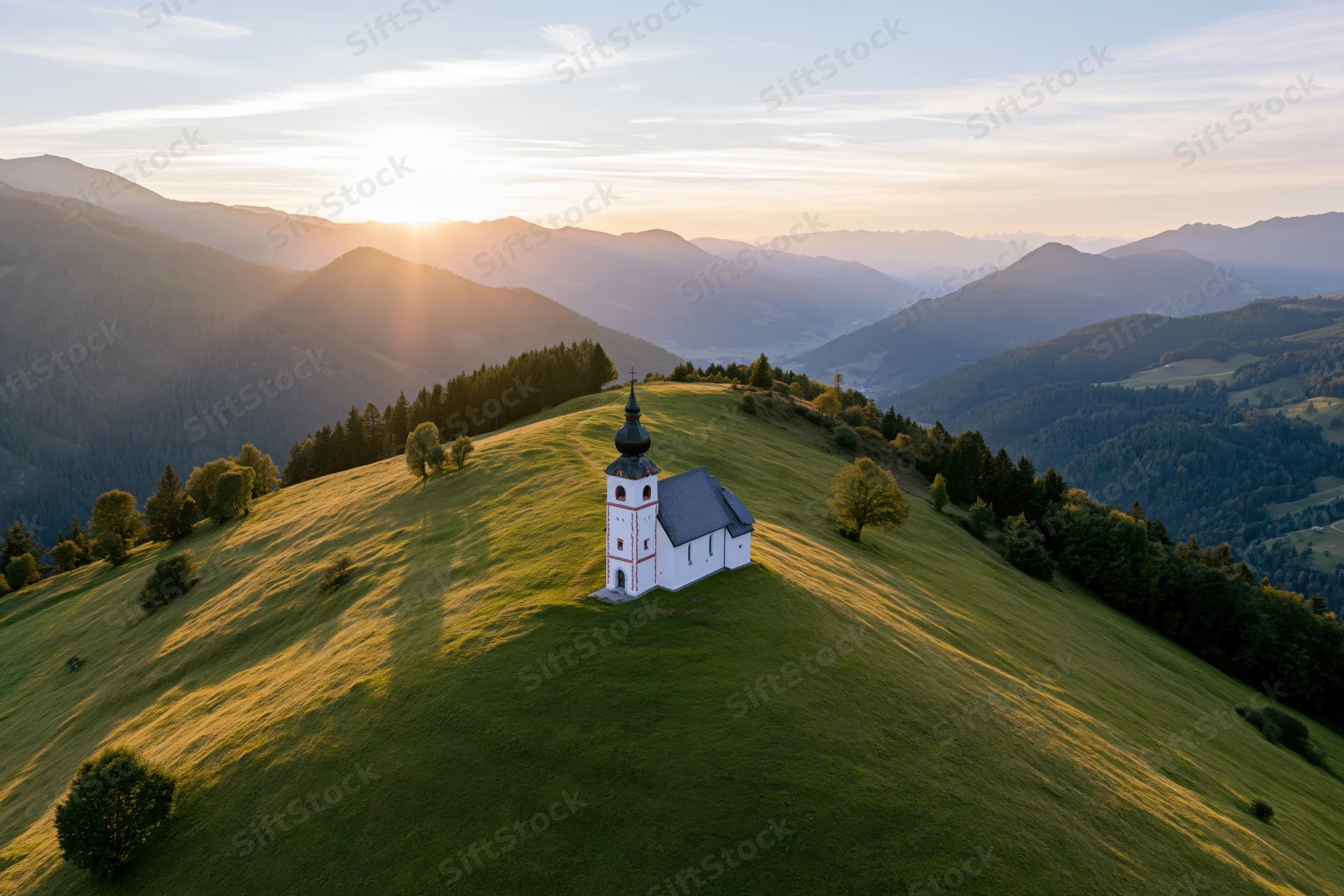 Small white alpine church on a grassy hill at sunset with mountains in the background