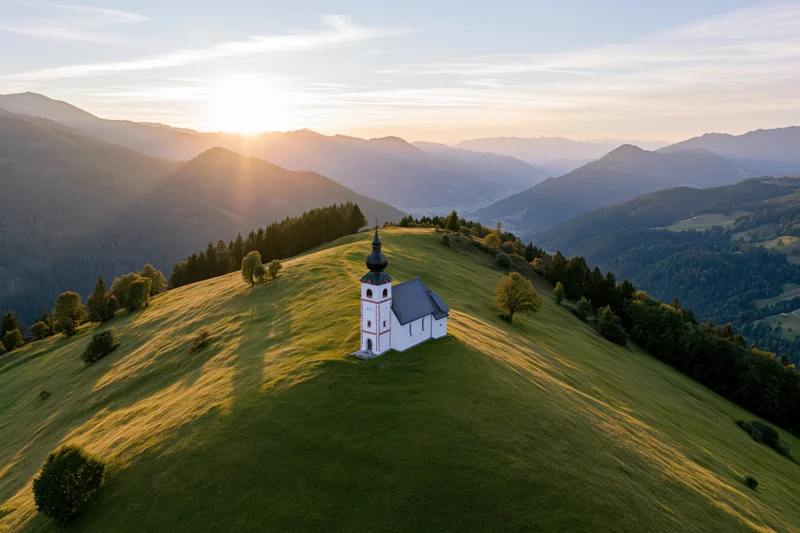 Small white alpine church on a grassy hill at sunset with mountains in the background