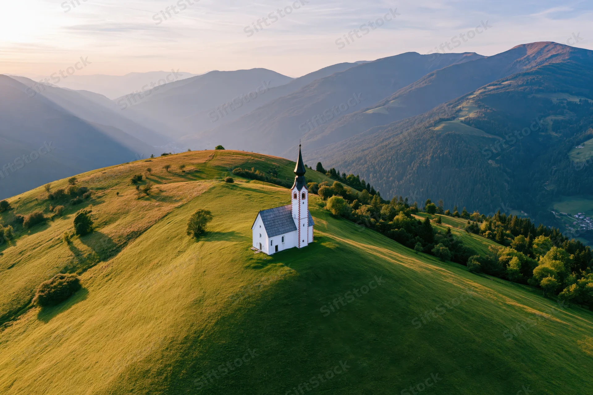 Small white alpine church on a grassy hilltop surrounded by mountains at sunset