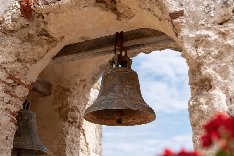 Aged bronze church bell hanging from wooden beam under stone arch with sky visible behind