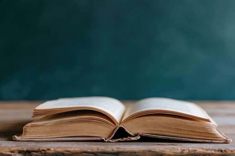 Open old hardcover book resting on worn wooden desk with teal background