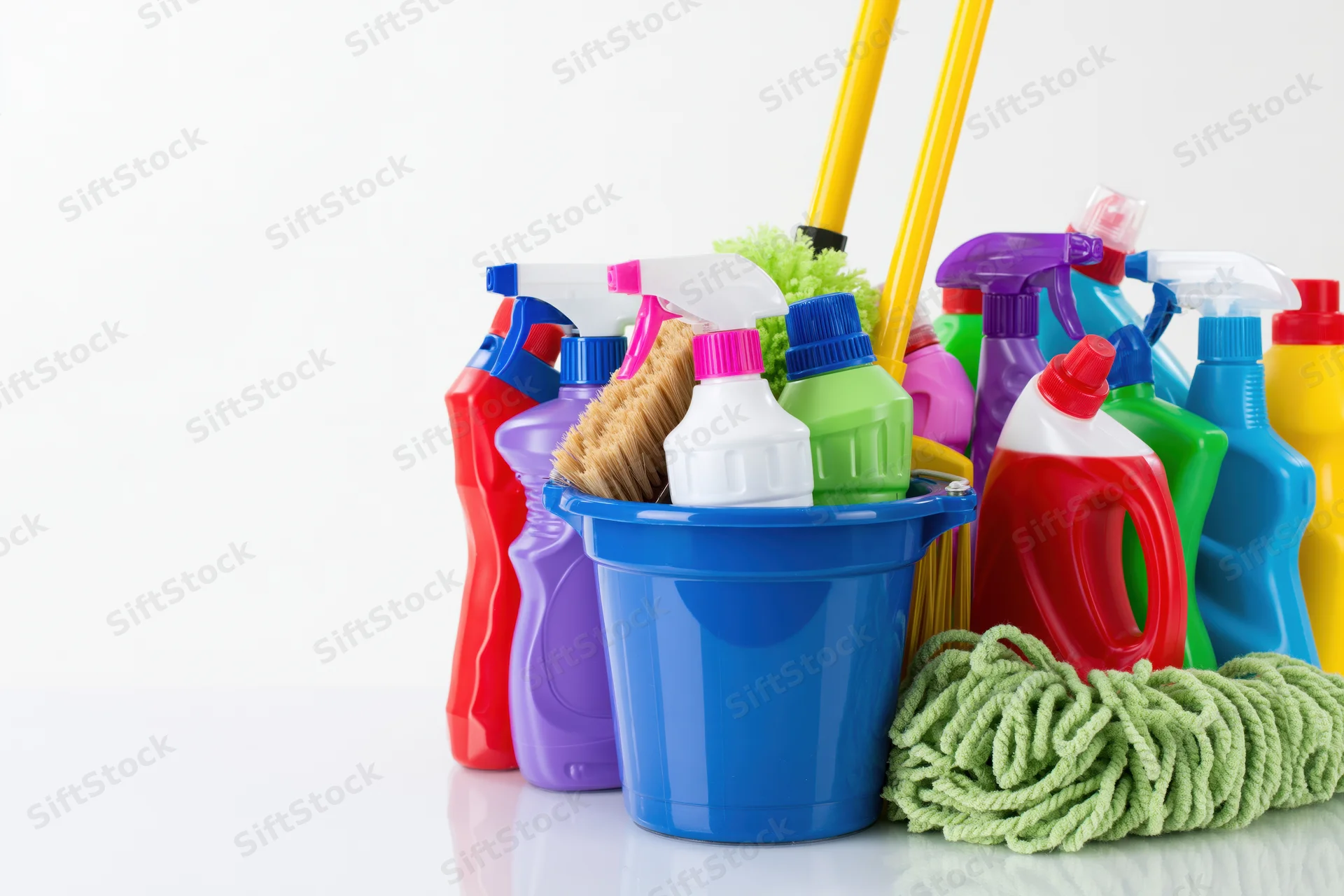 Assorted cleaning supplies arranged in a bucket with mops and bottles on a white surface