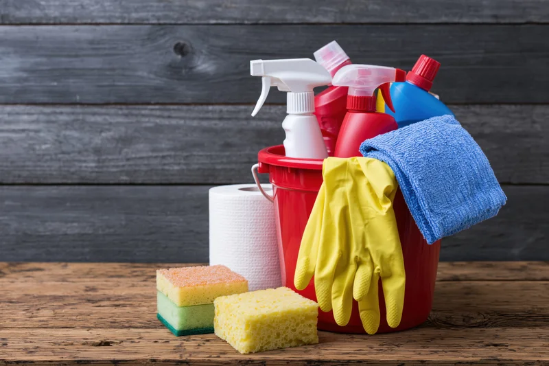 Cleaning supplies arranged in a red bucket on a wooden table with sponges, gloves and paper towels nearby