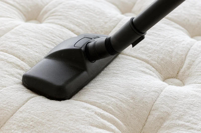 Black vacuum cleaner attachment cleaning a tufted white mattress surface