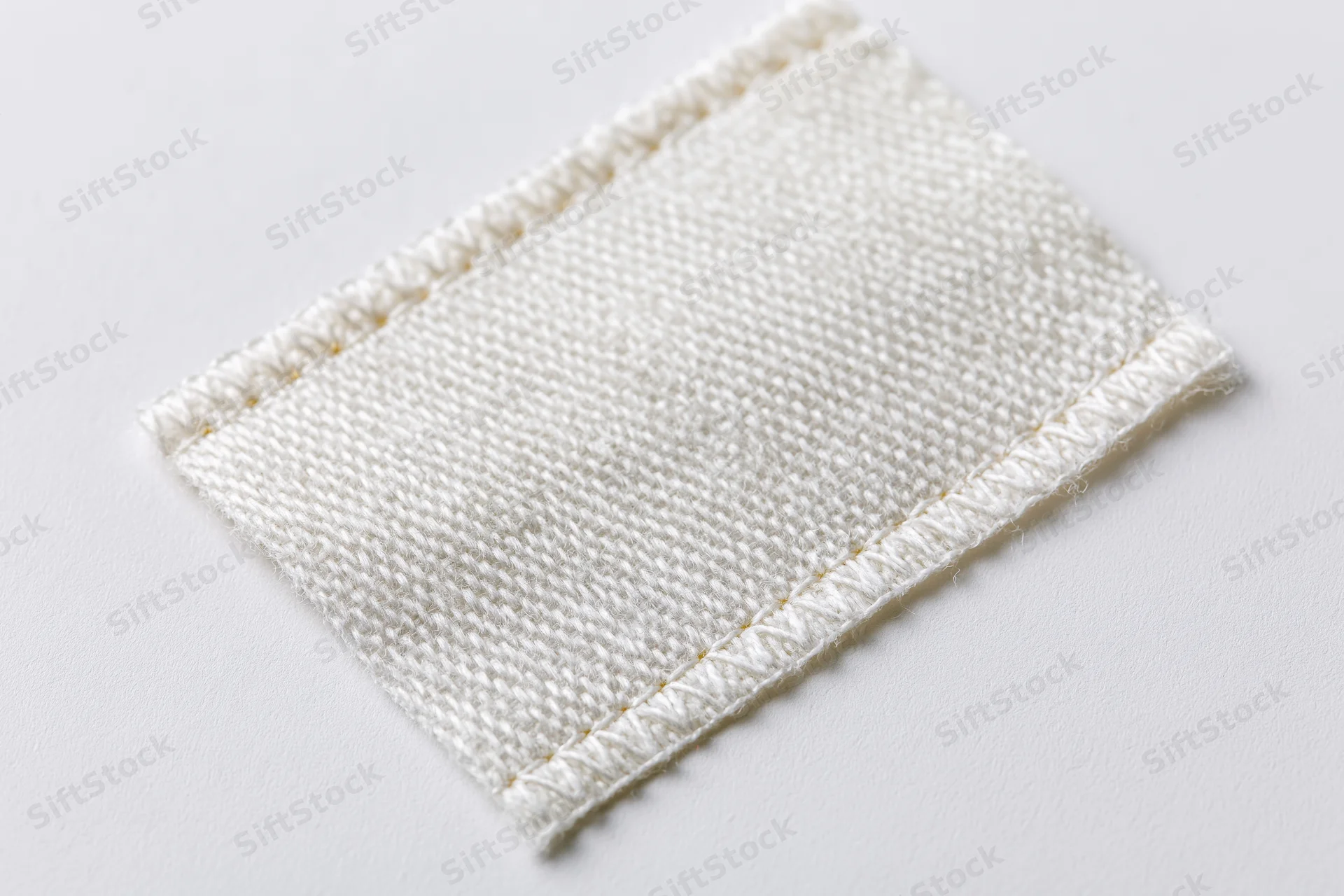 White rectangular fabric label stitched with thread lying flat on white surface