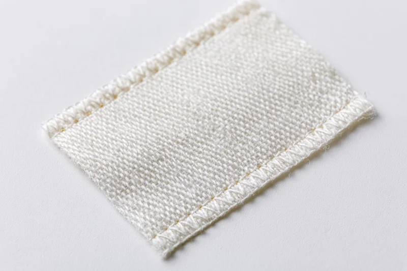White rectangular fabric label stitched with thread lying flat on white surface