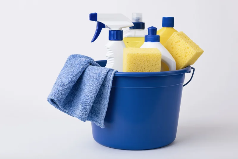 Blue plastic bucket filled with cleaning supplies including spray bottles, sponges and a towel on white background