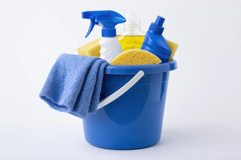 Blue plastic bucket filled with cleaning supplies including spray bottles, sponges, and a towel on white background