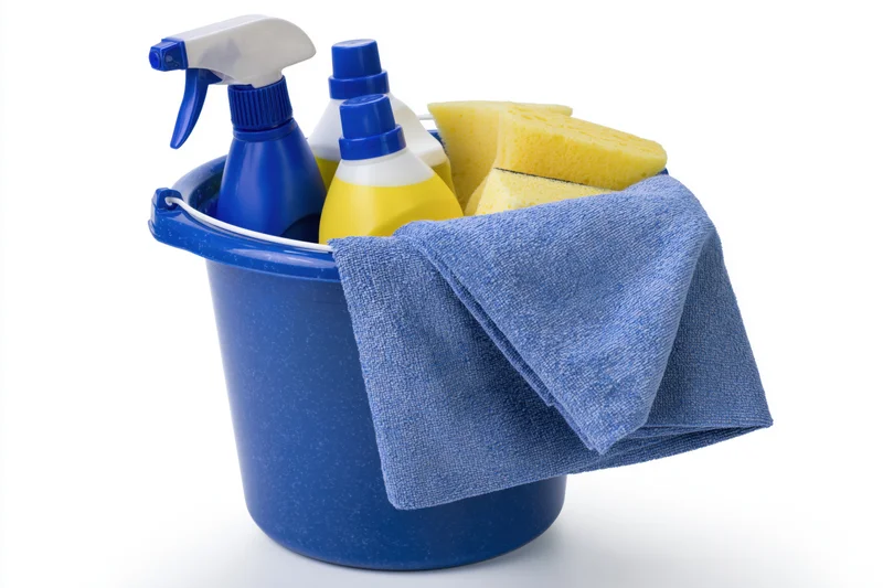 Blue plastic bucket filled with cleaning supplies including spray bottles, sponges and a towel on white background
