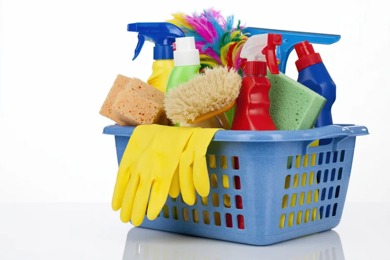 Blue plastic basket filled with cleaning supplies including spray bottles, sponges, gloves and brushes on white background
