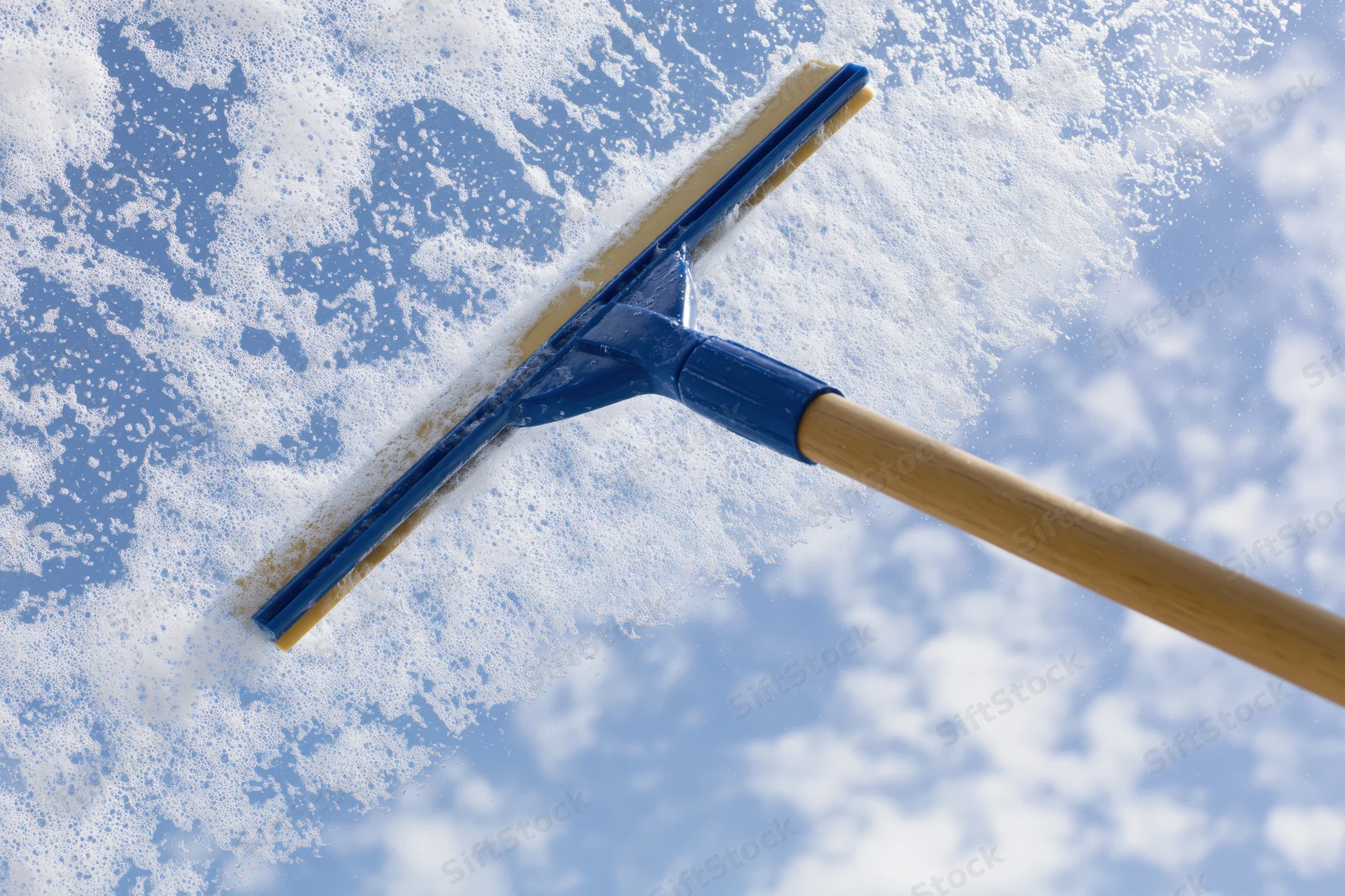 Blue rubber squeegee wiping soapy glass with sky visible through clean area