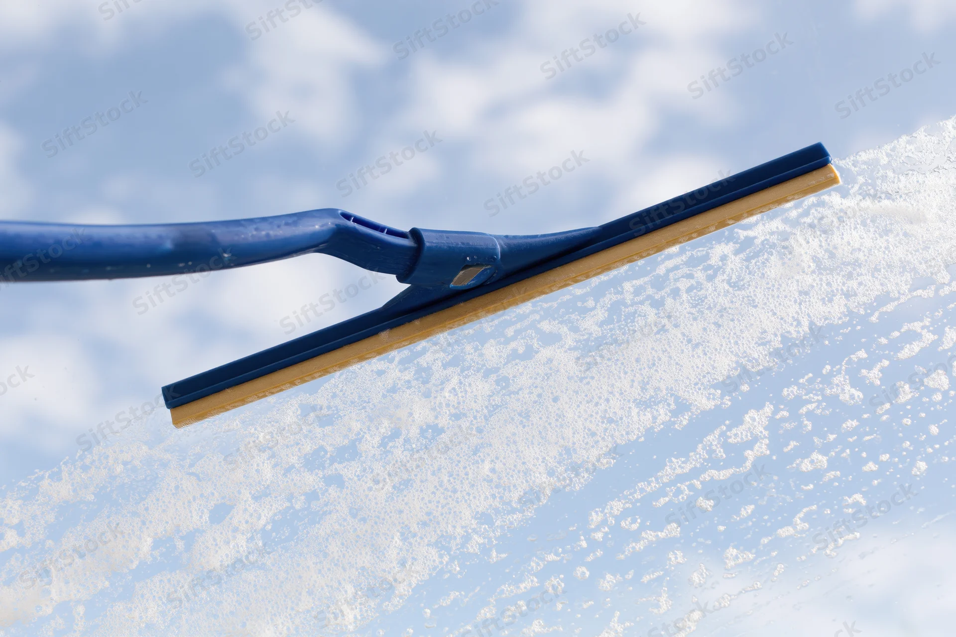 Blue rubber squeegee wiping soapy water from a glass window against a cloudy sky