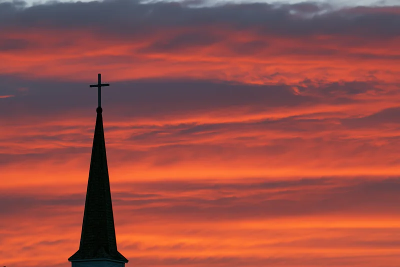 Church steeple with cross silhouetted against vibrant orange sunset sky