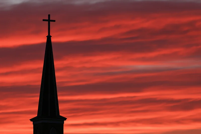 Church steeple with cross silhouetted against dramatic red sunset sky