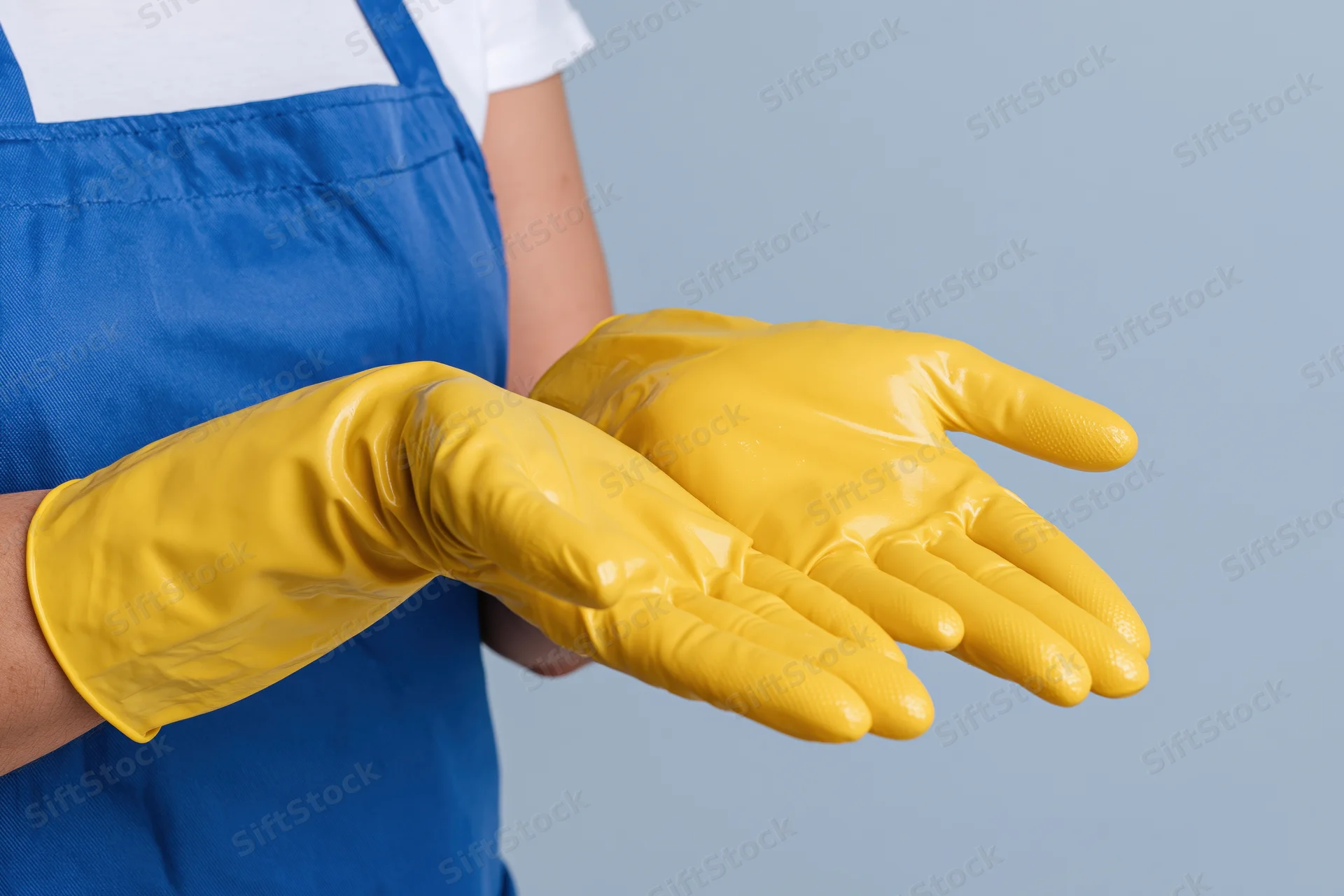 Woman in blue apron wearing yellow rubber gloves with hands extended against light blue background