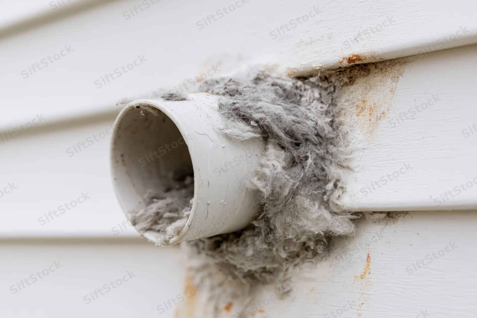 Clogged dryer vent pipe protruding from white vinyl siding with lint buildup