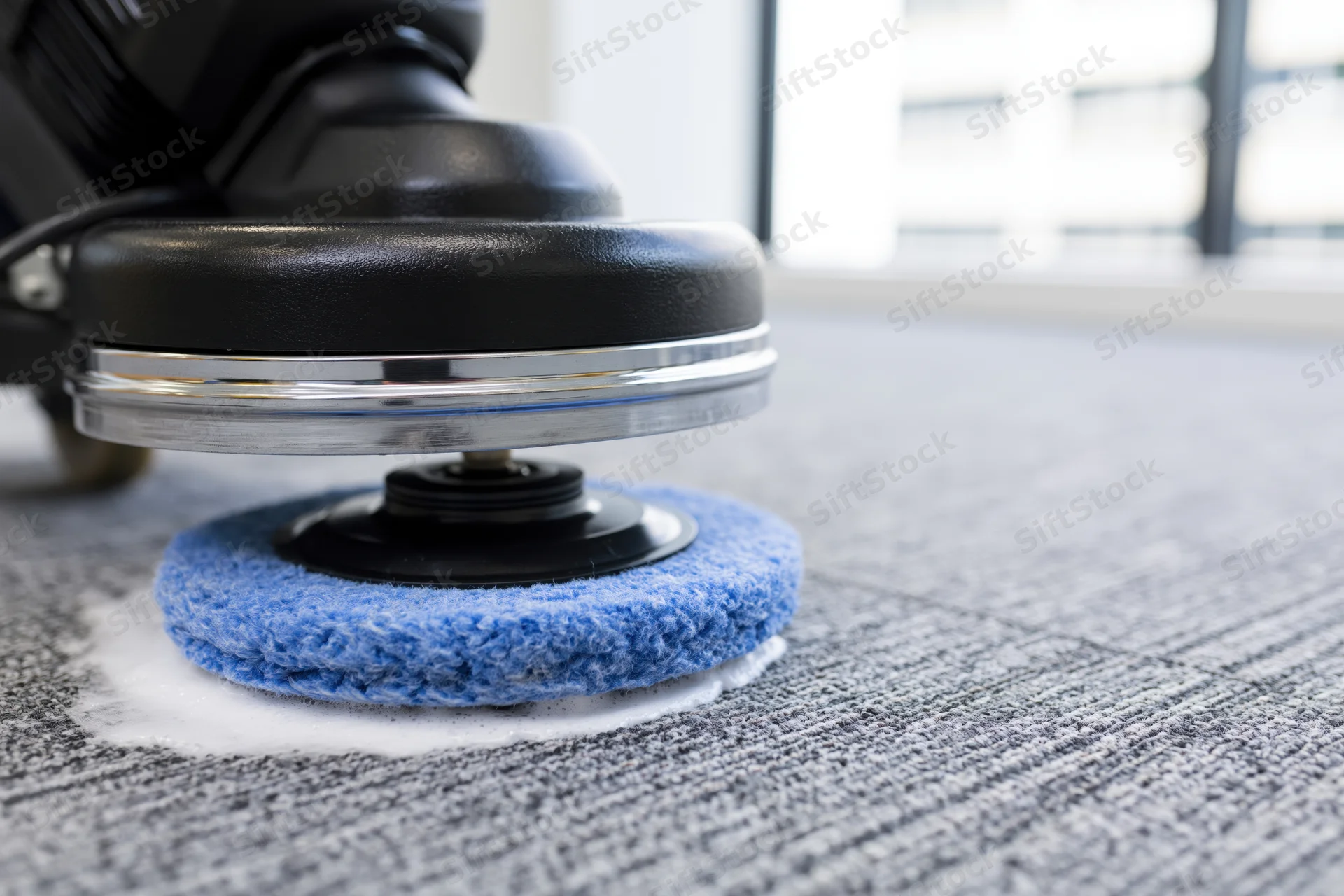 Commercial rotary floor polisher with blue scrubbing pad cleaning gray carpet in office interior