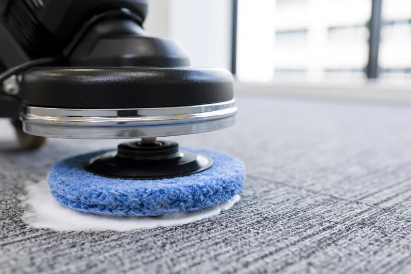 Commercial rotary floor polisher with blue scrubbing pad cleaning gray carpet in office interior