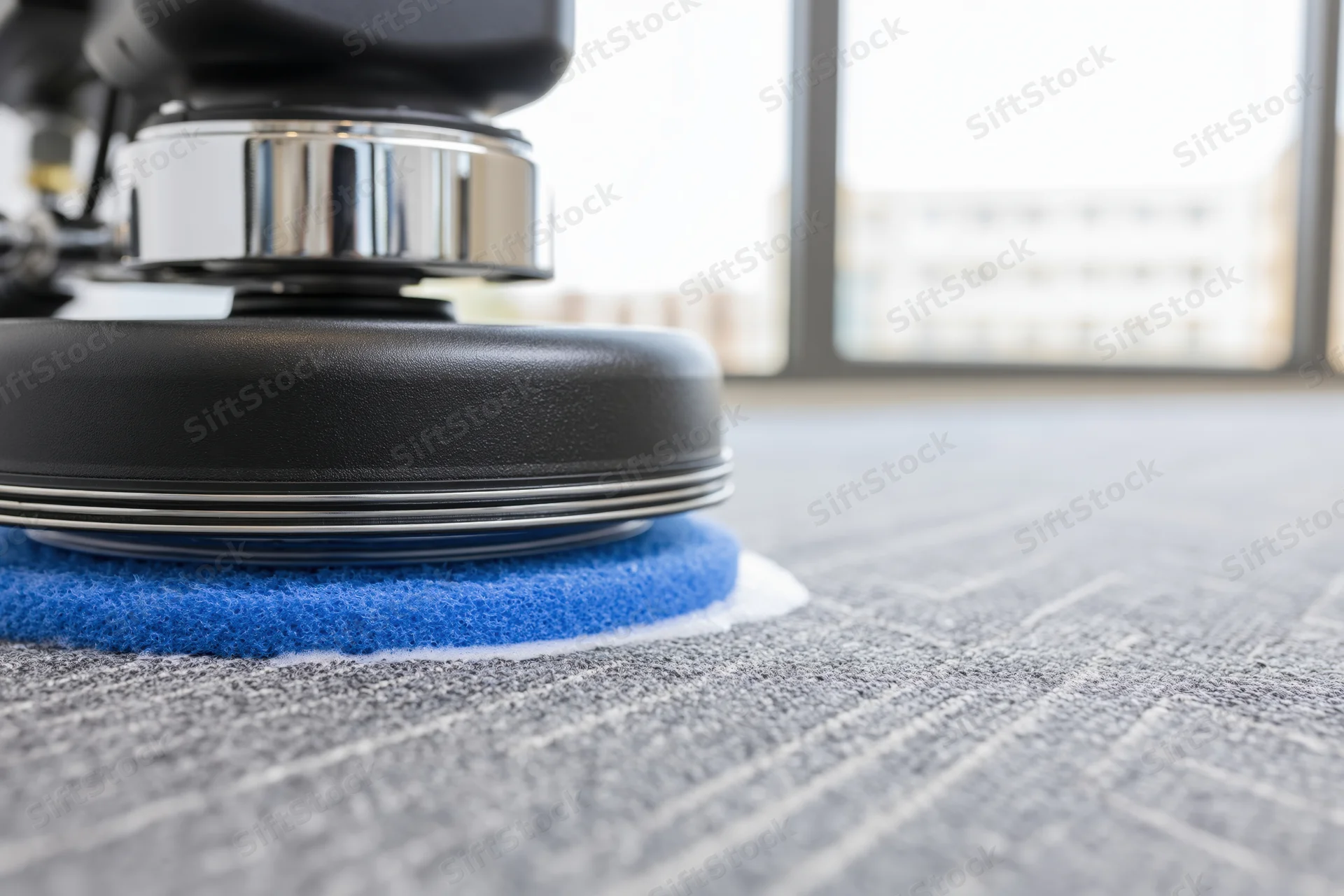 Commercial rotary floor polisher with blue scrubbing pad cleaning gray striped carpet in office interior