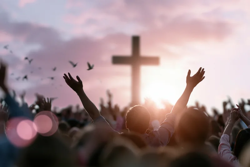 Silhouetted worshippers raising hands toward a large cross at sunset