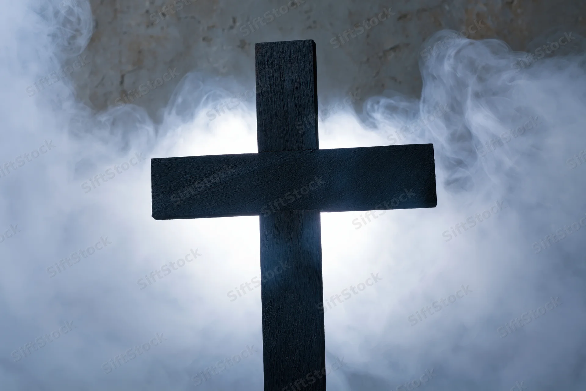 Dark wooden christian cross silhouetted against swirling smoke with a glowing light behind it