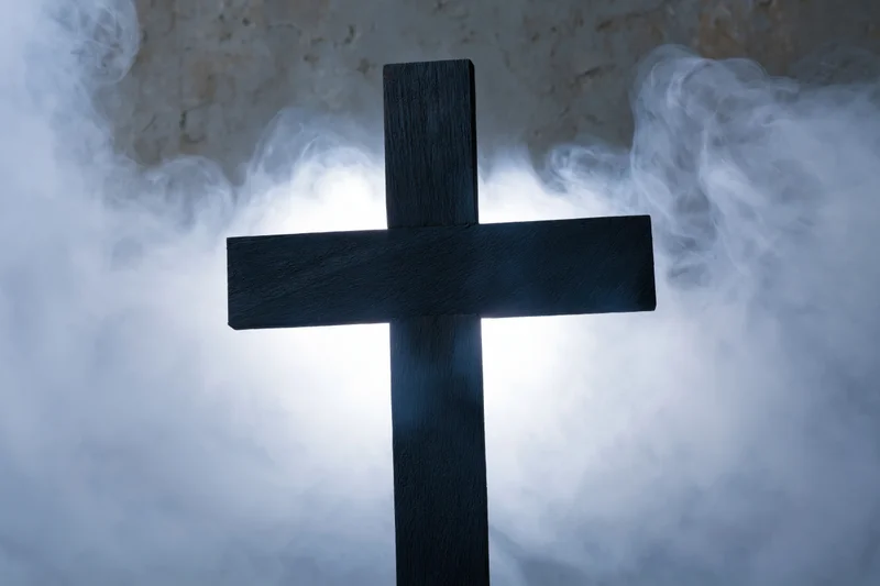 Dark wooden christian cross silhouetted against swirling smoke with a glowing light behind it