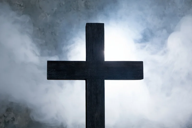 Dark wooden christian cross silhouetted against smoky sky with light breaking through