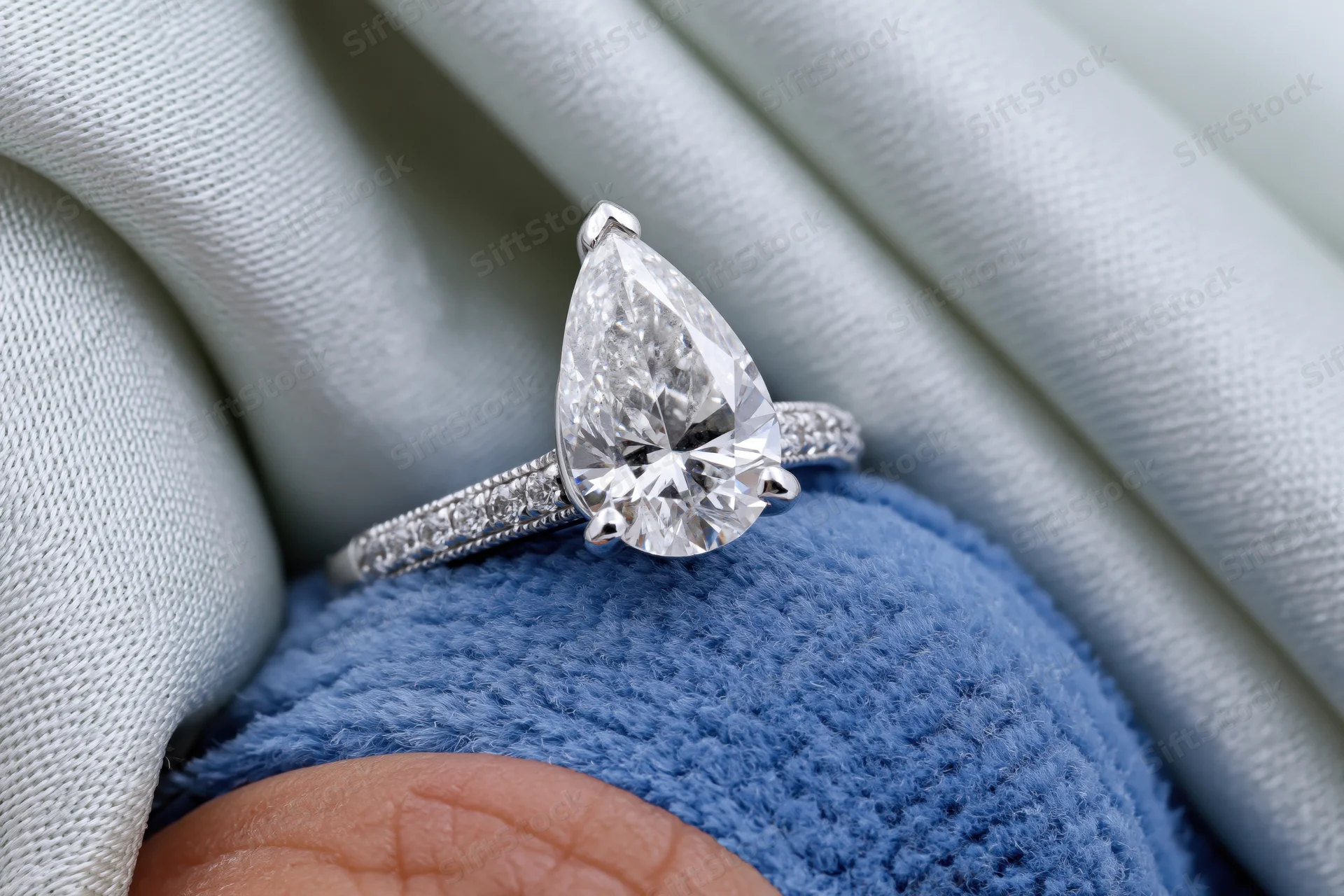 Pear-cut diamond solitaire engagement ring resting on a blue knitted fabric held by a hand