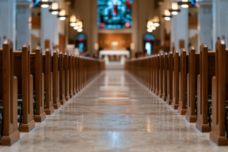 View down the central aisle of a Catholic church nave lined with wooden pews toward the altar and stained glass window