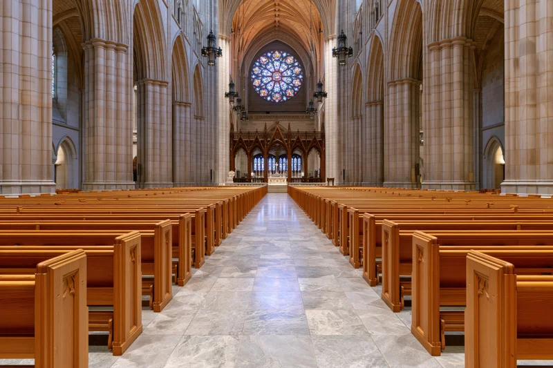Central aisle of a gothic cathedral nave lined with wooden pews leading to a stained glass rose window and altar