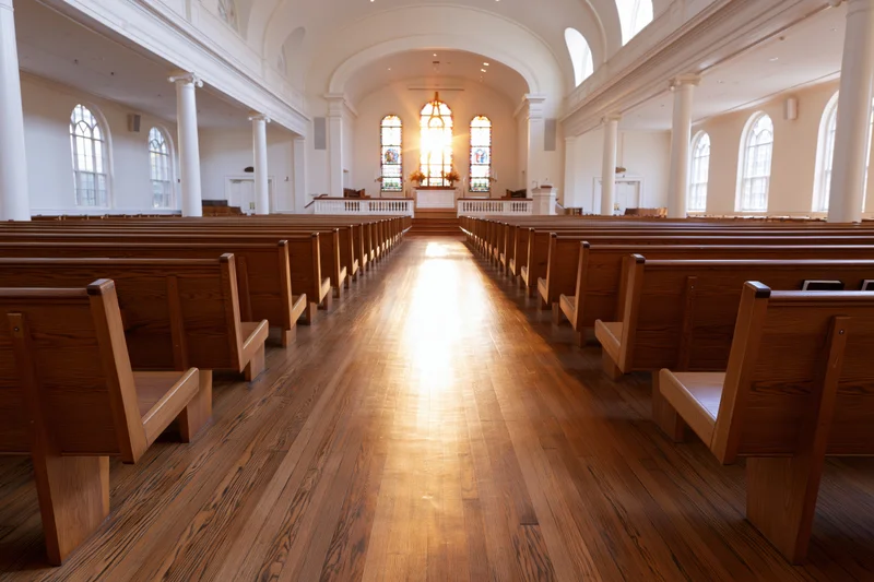Empty church interior viewed down central aisle toward altar with sunlight streaming through stained glass windows