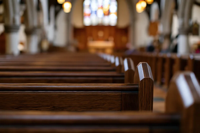 Rows of wooden pews lining the central aisle in a church interior with stained glass and altar in the distance