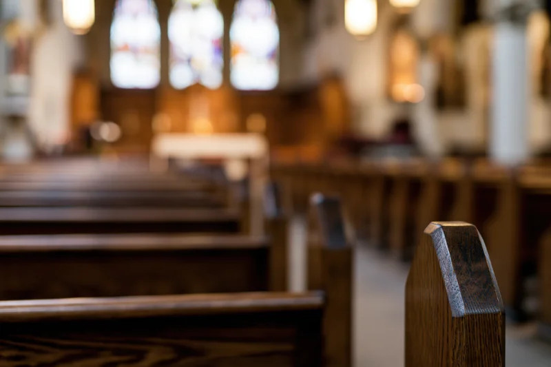 Wooden pews lining the central aisle in a church interior with stained glass windows in the background