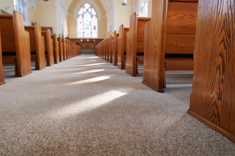 Rows of wooden pews lining a carpeted aisle in a church interior with sunlight streaming through stained glass windows