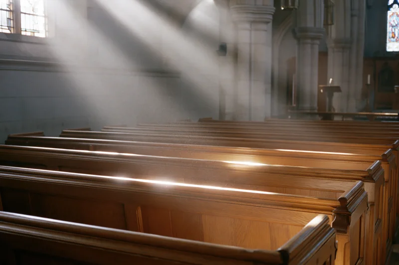 Rows of wooden pews in a church interior illuminated by sunlight streaming through stained glass windows