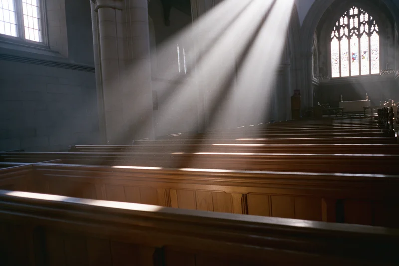 Rows of wooden pews in a church interior with sunlight streaming through stained glass windows