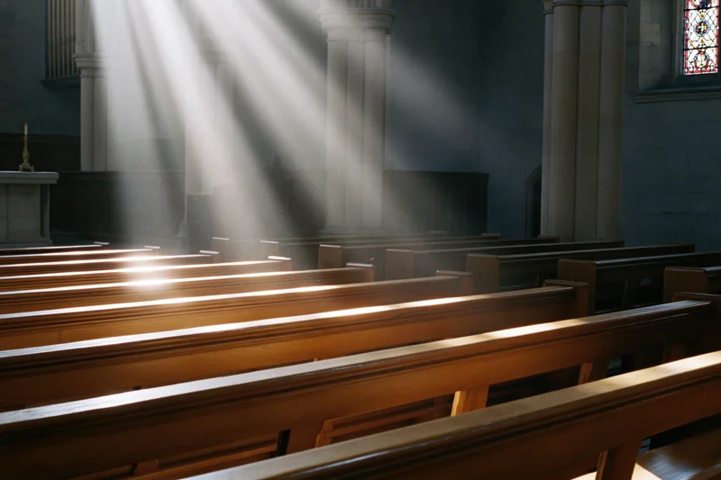 Rows of wooden pews in a church interior illuminated by sunlight streaming through stained glass windows