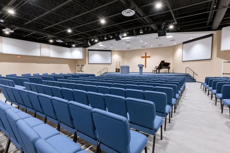 Rows of blue padded chairs in a contemporary church sanctuary facing a stage with podium, cross, and piano