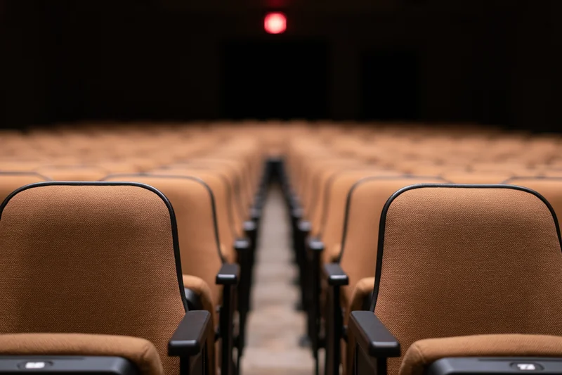 Rows of empty upholstered theater seats stretching toward a dim stage with a red exit light at the end