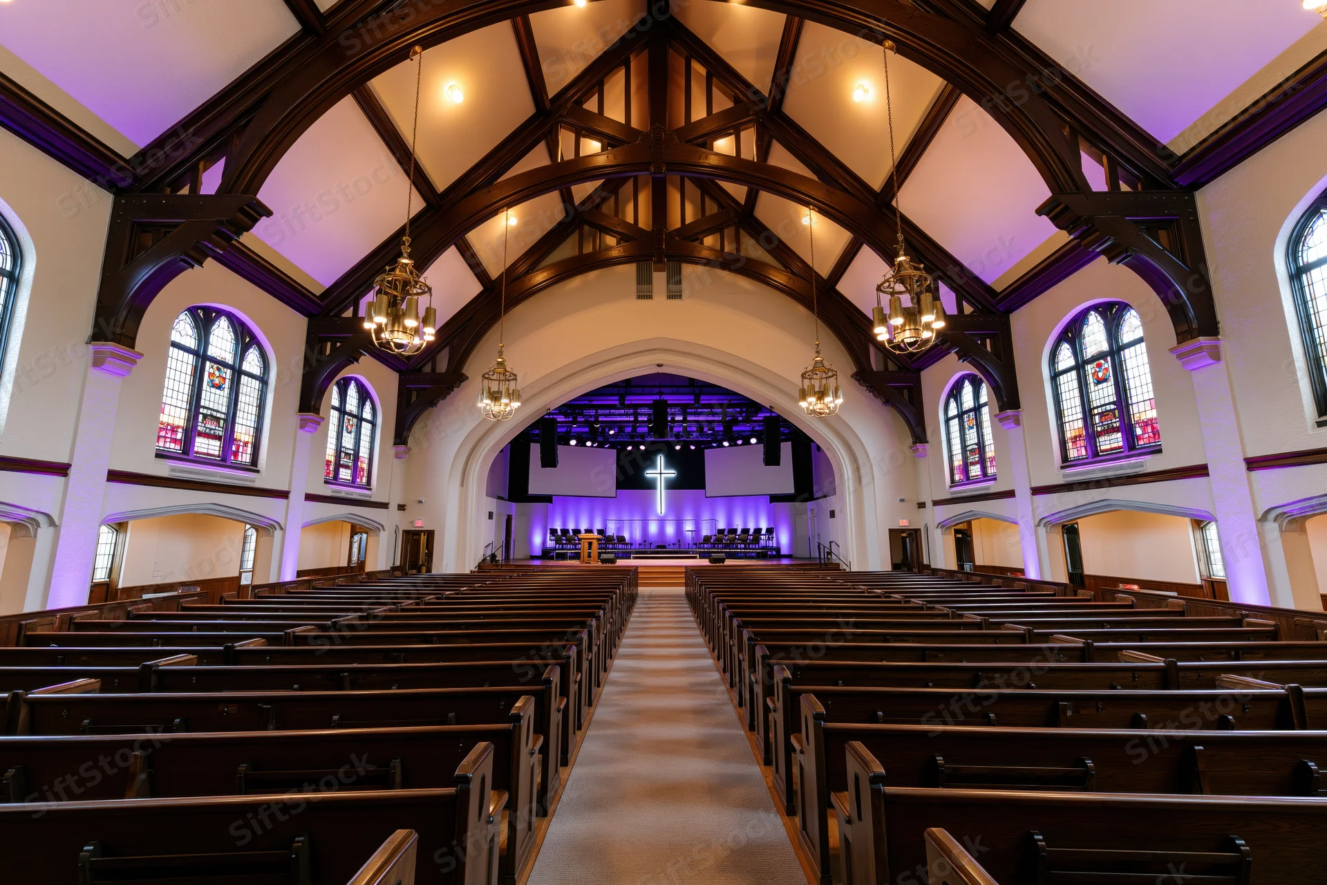 Empty traditional church sanctuary interior viewed from the back with wooden pews, arched ceiling, stained glass windows, and purple-lit altar at front