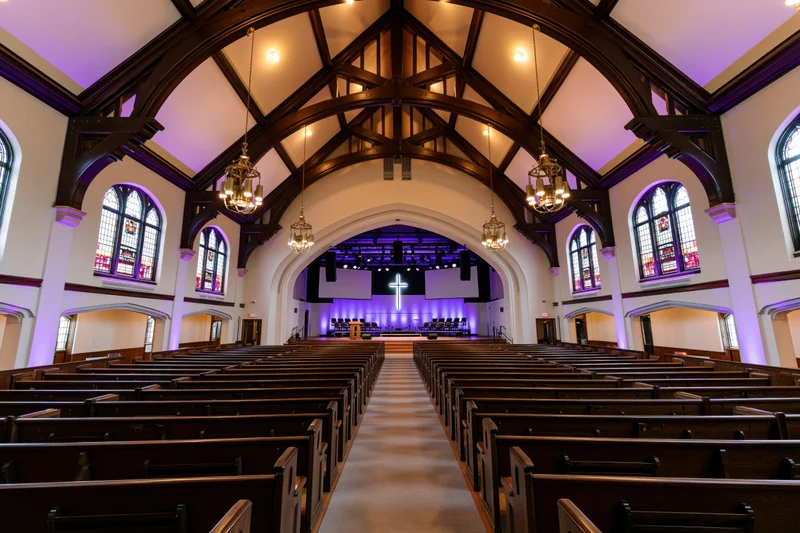 Empty traditional church sanctuary interior viewed from the back with wooden pews, arched ceiling, stained glass windows, and purple-lit altar at front